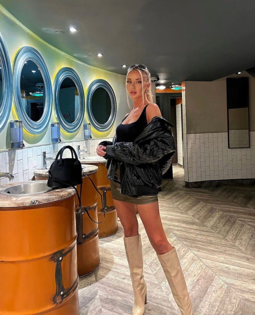 Woman in black jacket and boots in bathroom.